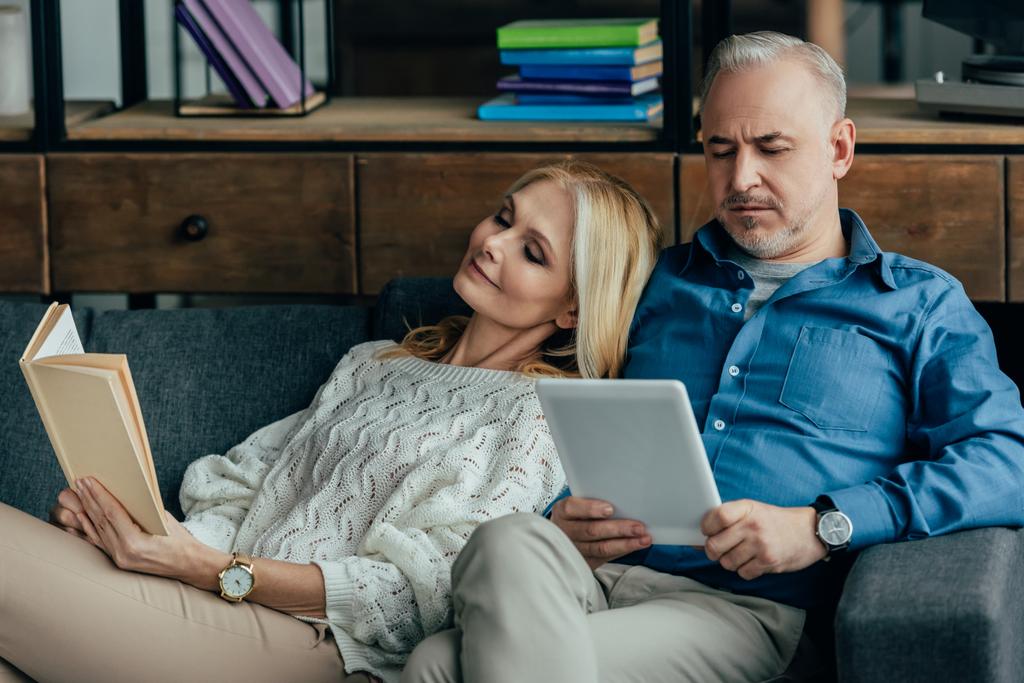feliz esposa leyendo libro cerca de marido usando tableta digital mientras está sentado en sofá
 - Foto, imagen