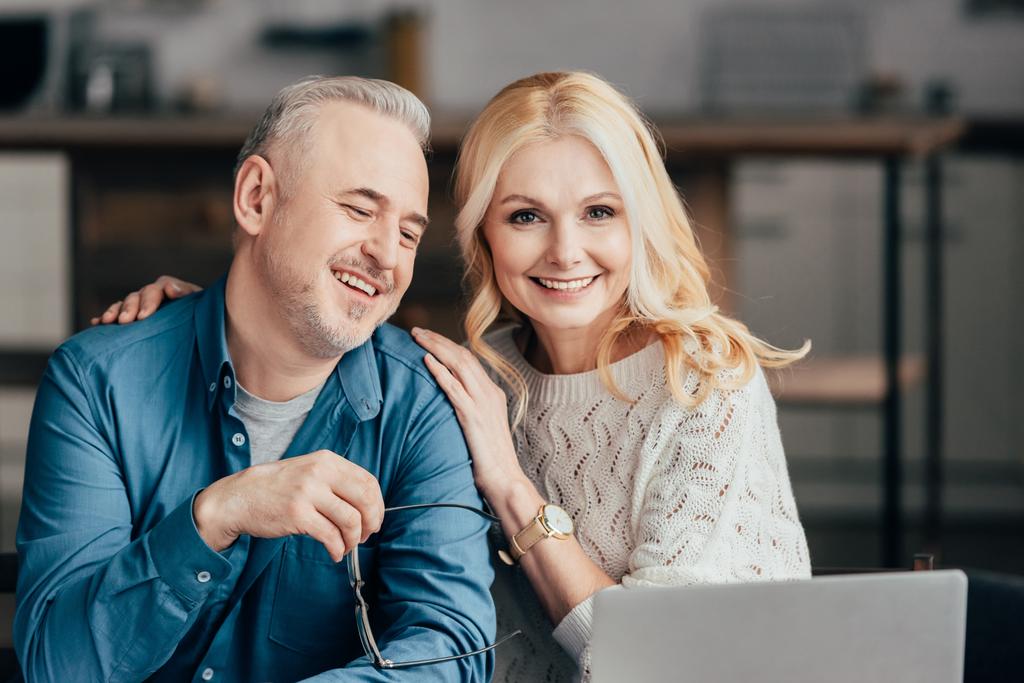 homem bonito segurando óculos e sorrindo com esposa alegre enquanto olha para laptop
 - Foto, Imagem