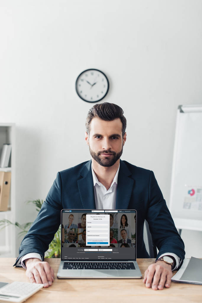 όμορφος σύμβουλος στο κοστούμι δείχνει laptop με linkedin ιστοσελίδα στην οθόνη στο γραφείο  - Φωτογραφία, εικόνα