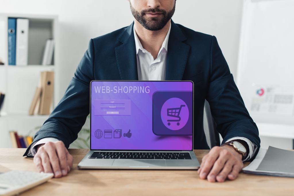 vista ritagliata del consulente in giacca e cravatta che mostra laptop con sito web-shopping sullo schermo in ufficio
  - Foto, immagini