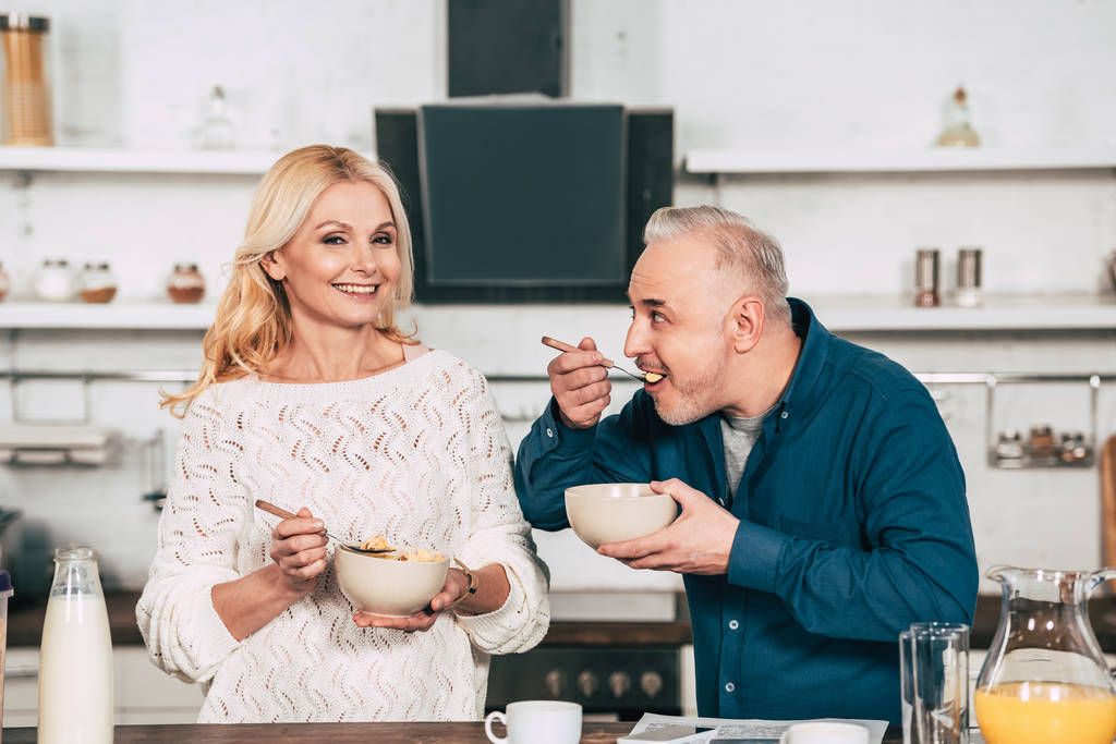 attractive wife holding bowl with cornflakes near cheerful husband eating in kitchen - Photo, Image