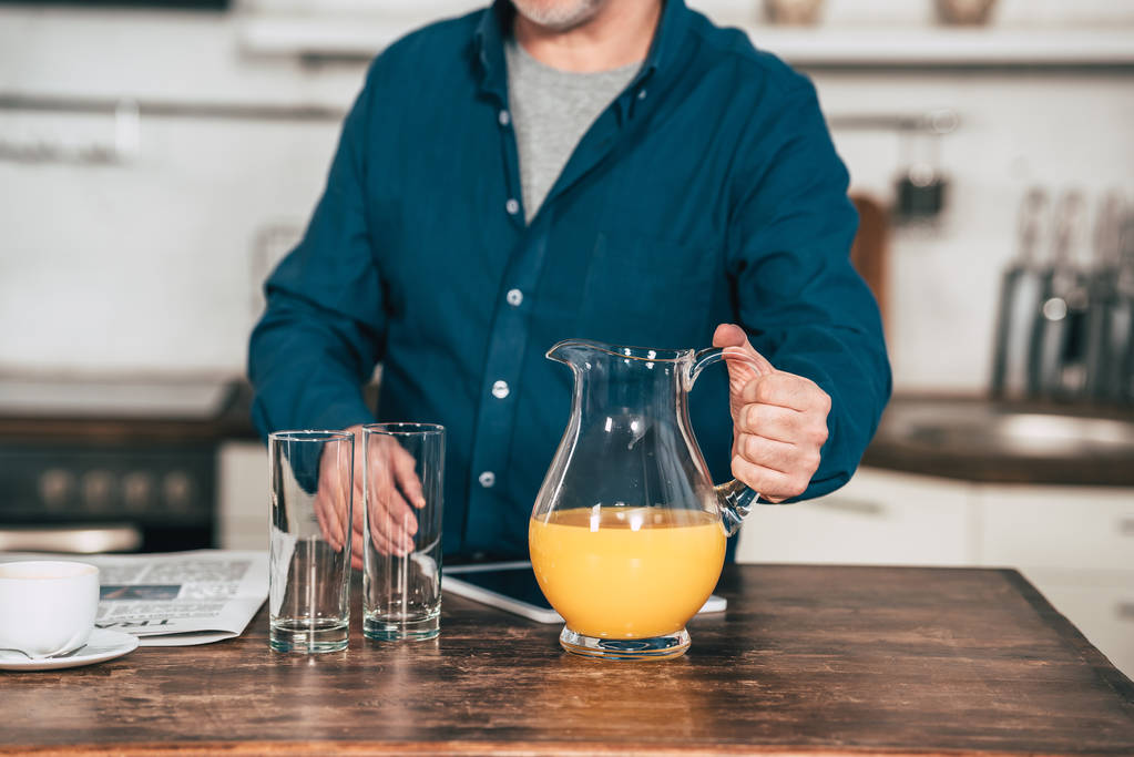 περικοπεί θεώρηση του ανθρώπου που κρατώντας την κανάτα με χυμό πορτοκαλιού κοντά γυαλιά στην κουζίνα - Φωτογραφία, εικόνα