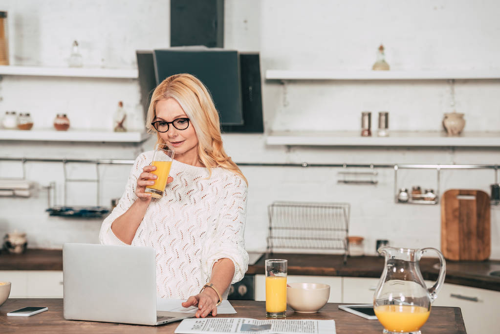 atractiva mujer en gafas para los ojos beber jugo de naranja cerca de la computadora portátil
 - Foto, Imagen