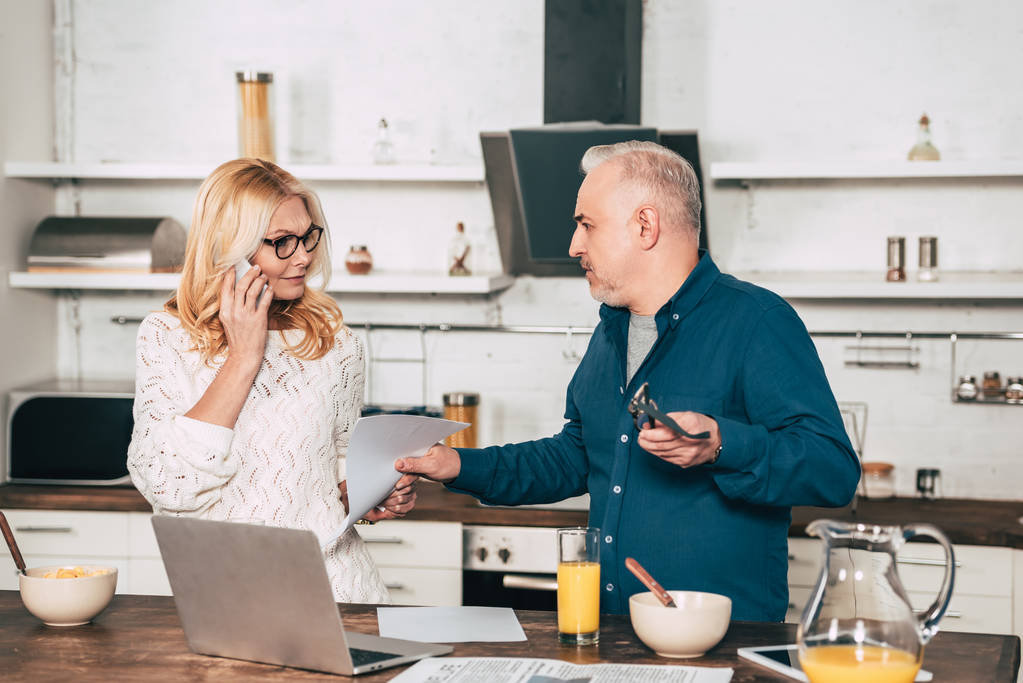atractiva mujer hablando en el teléfono inteligente y de pie con el marido en la cocina cerca de la computadora portátil
 - Foto, imagen