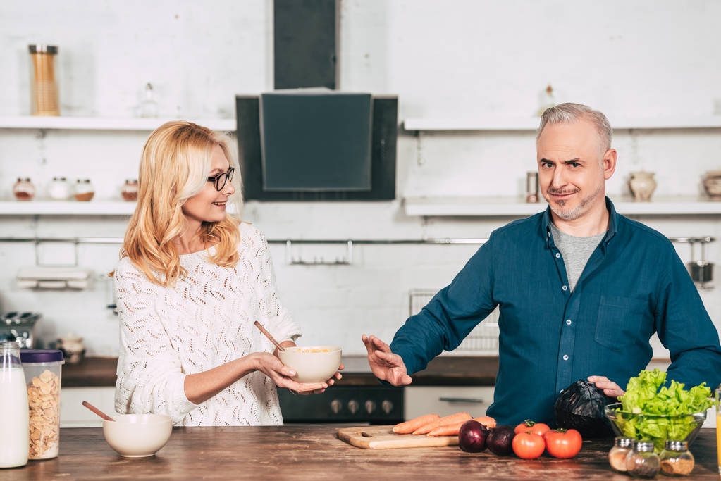 attraktive Frau in Gläsern mit Schale neben Mann gestikulierend in Küche - Foto, Bild