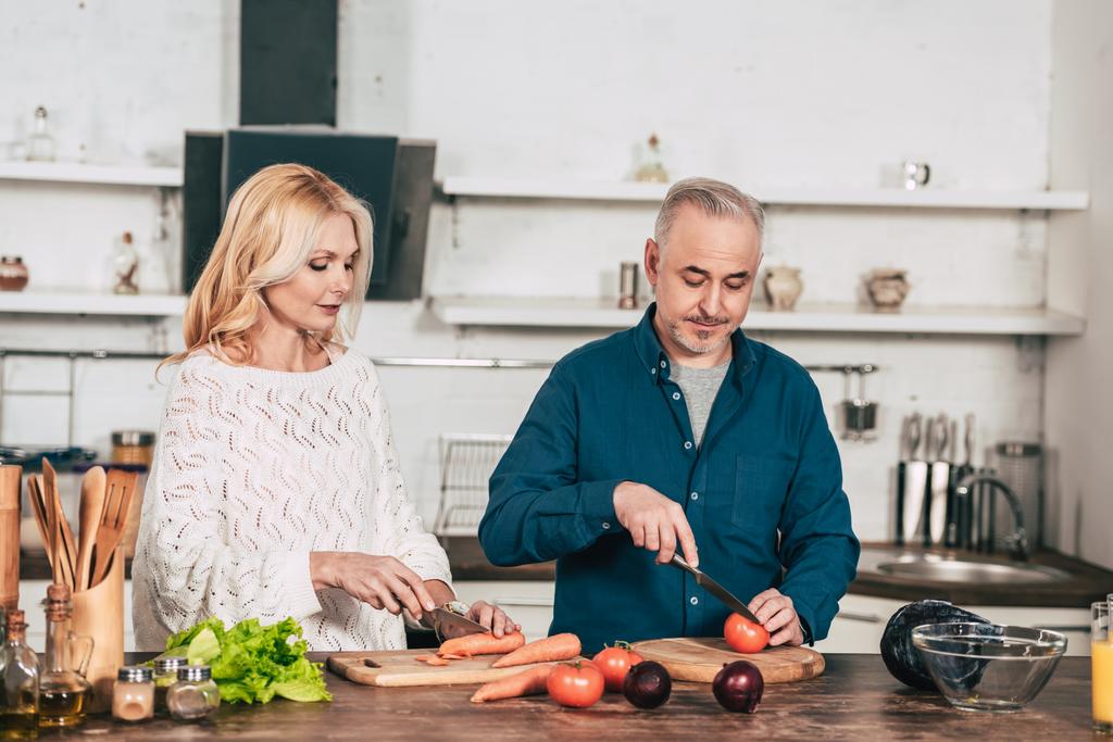attraktive Frau schneidet Karotte in der Nähe Ehemann hält Messer in der Nähe Tomate in der Küche - Foto, Bild