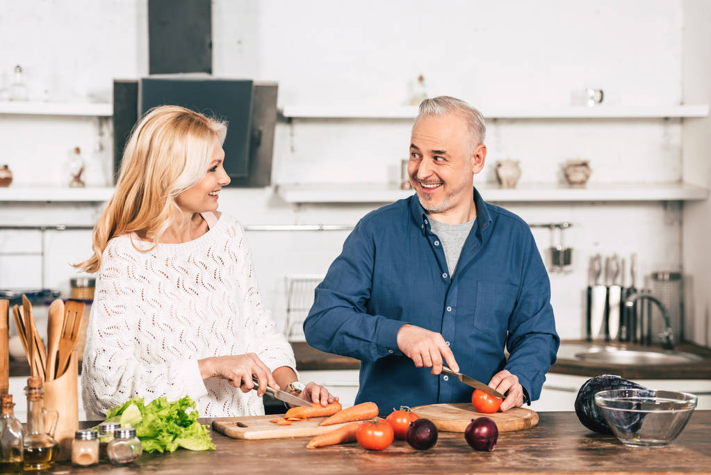 cheerful couple cutting carrot and tomato and smiling in kitchen - Photo, Image
