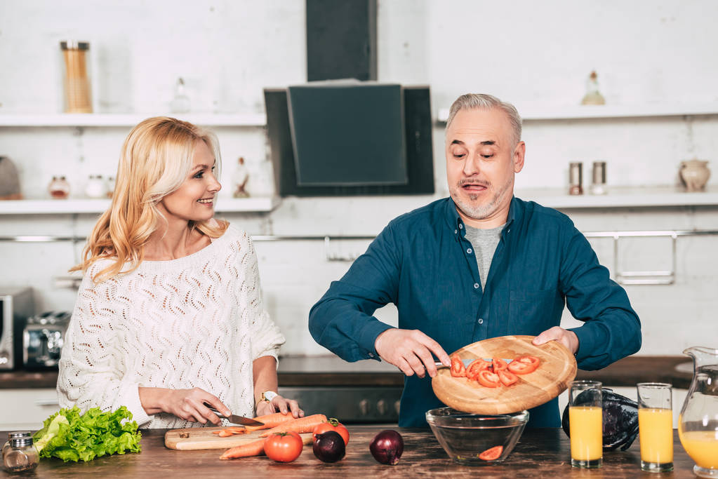 mujer alegre cortando zanahoria y mirando marido poniendo tomates cortados en tazón de cristal
   - Foto, Imagen