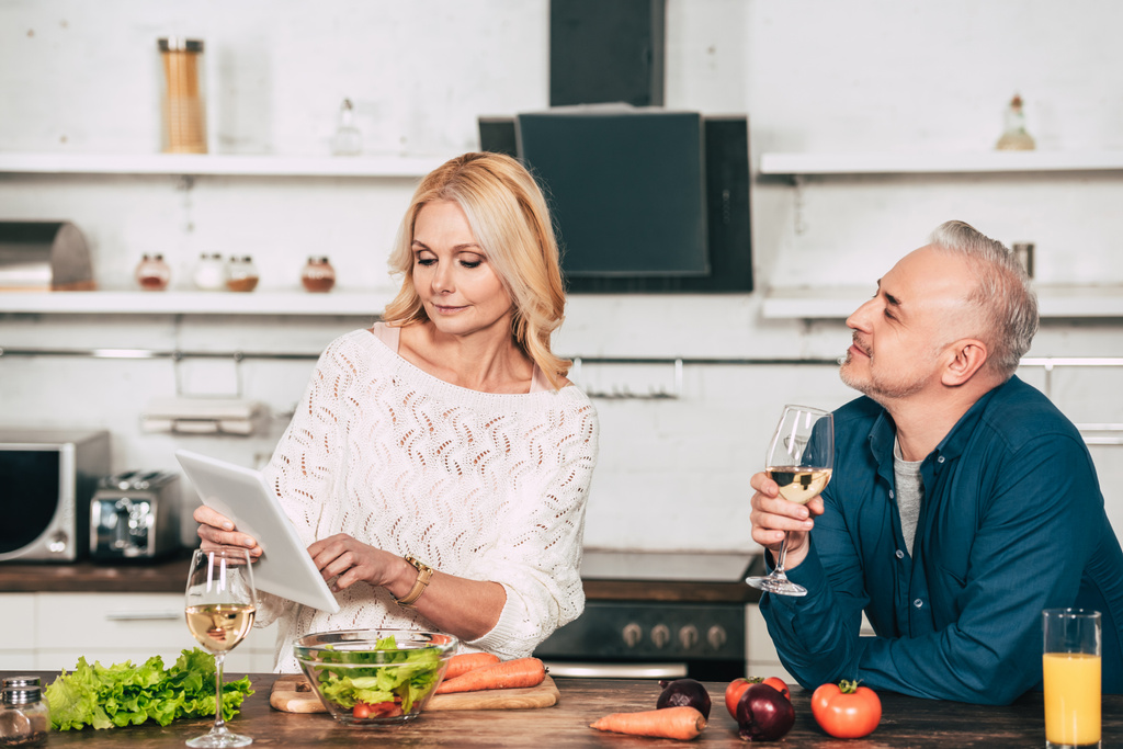 όμορφος άνδρας κρατώντας το ποτήρι με το κρασί και να εξετάσουμε ελκυστική ξανθιά γυναίκα κρατώντας ψηφιακό tablet στην κουζίνα - Φωτογραφία, εικόνα