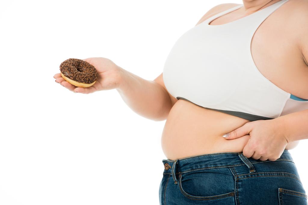 bijgesneden weergave van overgewicht vrouw houden van zoete donut en aanraken van de buik geïsoleerd op wit, concept dieet - Foto, afbeelding