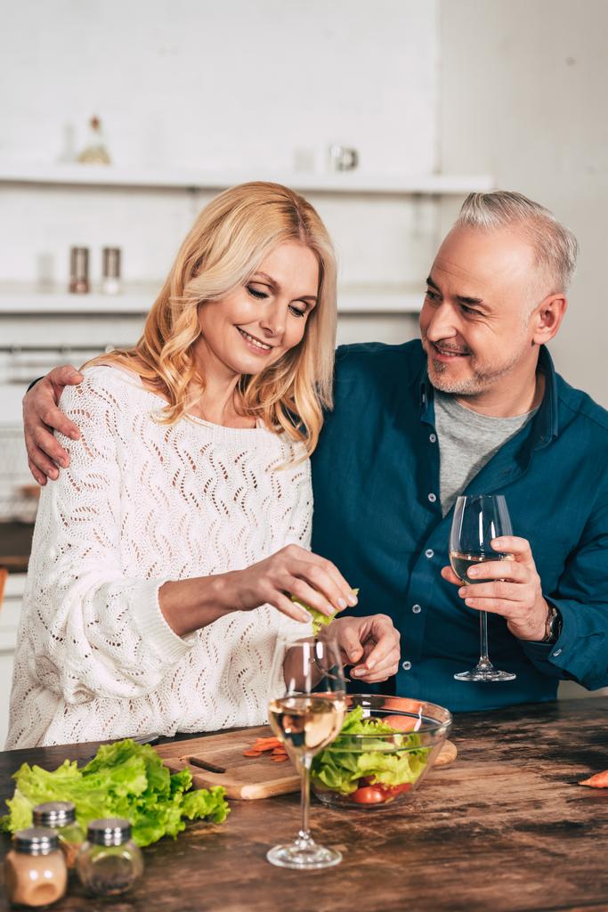 schöner Mann mit einem Glas Wein umarmt attraktive blonde Frau mit grünen Salatblättern - Foto, Bild
