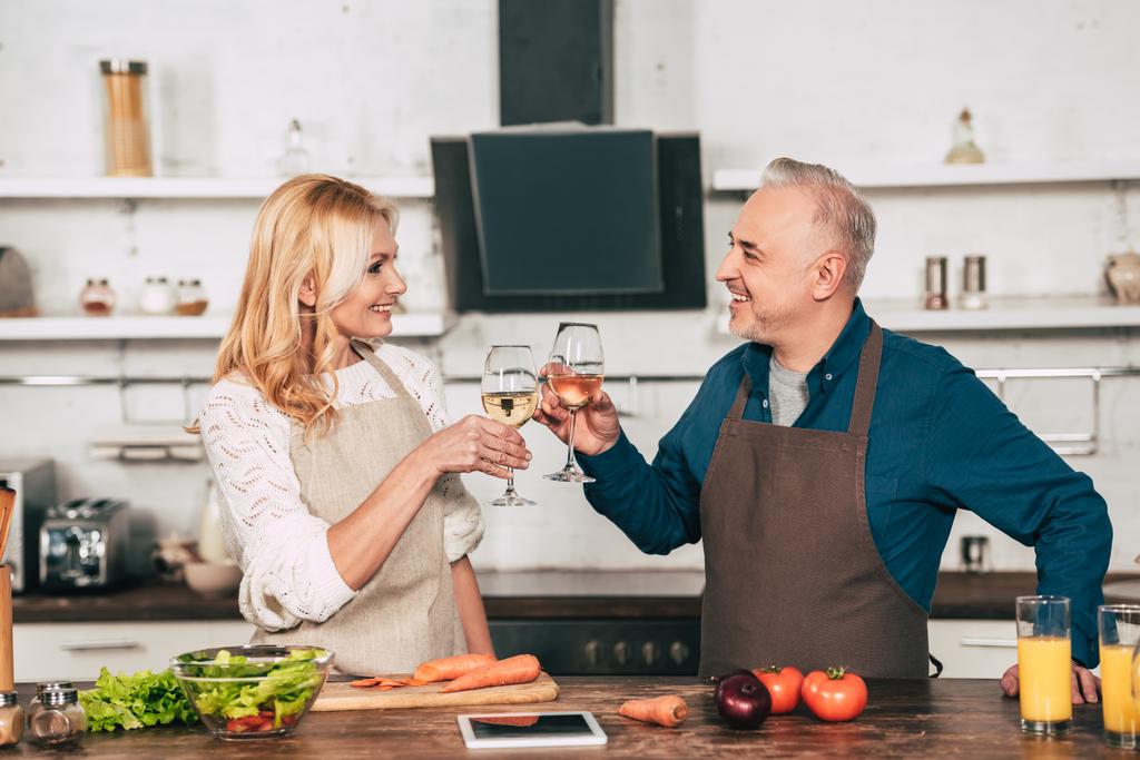 pohledný manžel cinkání sklenic vína s usměvavou manželku v kuchyni - Fotografie, Obrázek