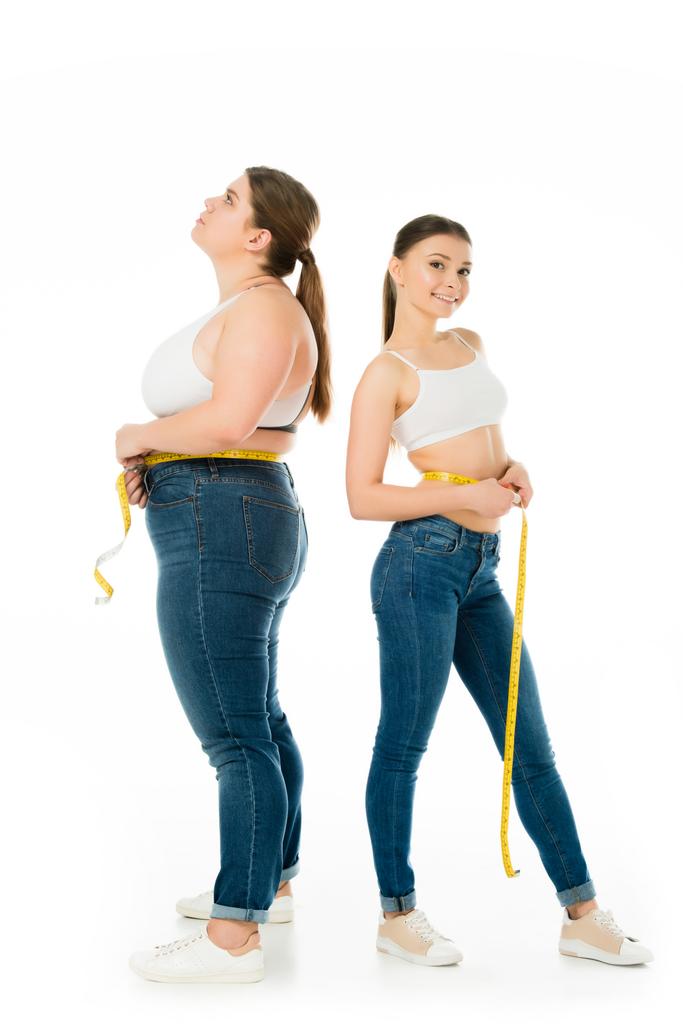 triste donna in sovrappeso con metro a nastro guardando verso l'alto mentre attraente donna magra guardando la fotocamera isolata su bianco
 - Foto, immagini