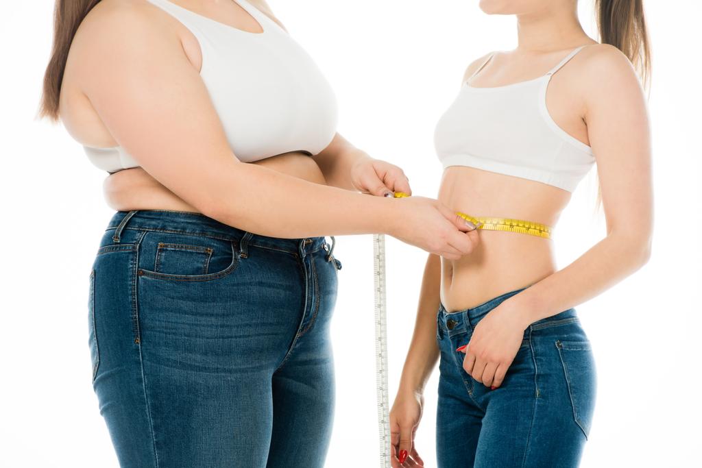 abgeschnittene Ansicht einer übergewichtigen Frau, die die Taille einer schlanken Frau misst, isoliert auf Weiß - Foto, Bild