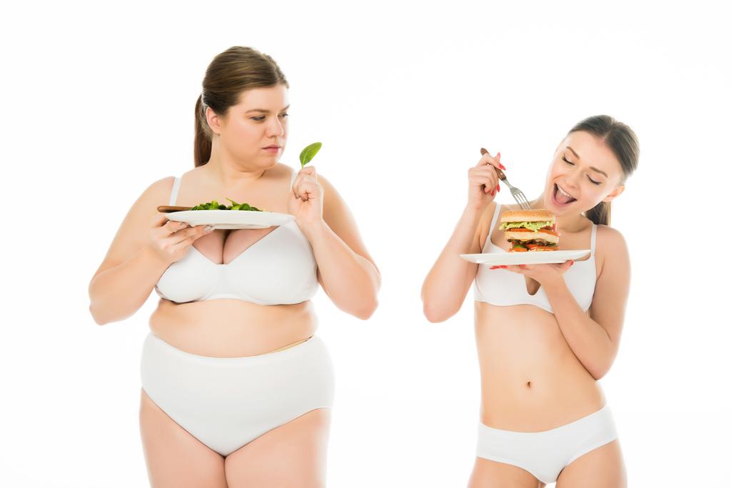 スリムな女性下着の太りすぎは、緑のほうれん草を保持している女性を混乱させる間、ハンバーガーを食べることで葉に分離ホワイト - 写真・画像
