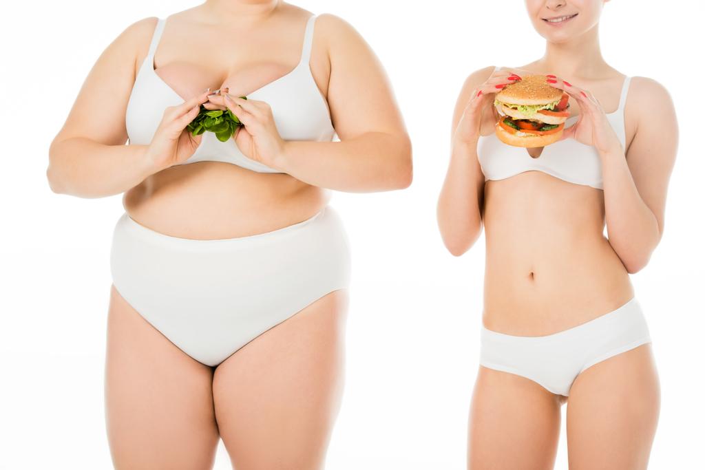 szczupła kobieta uśmiechający się w bieliźnie, trzymając burger podczas nadwagę kobieta trzyma zielony szpinak liście na białym tle - Zdjęcie, obraz