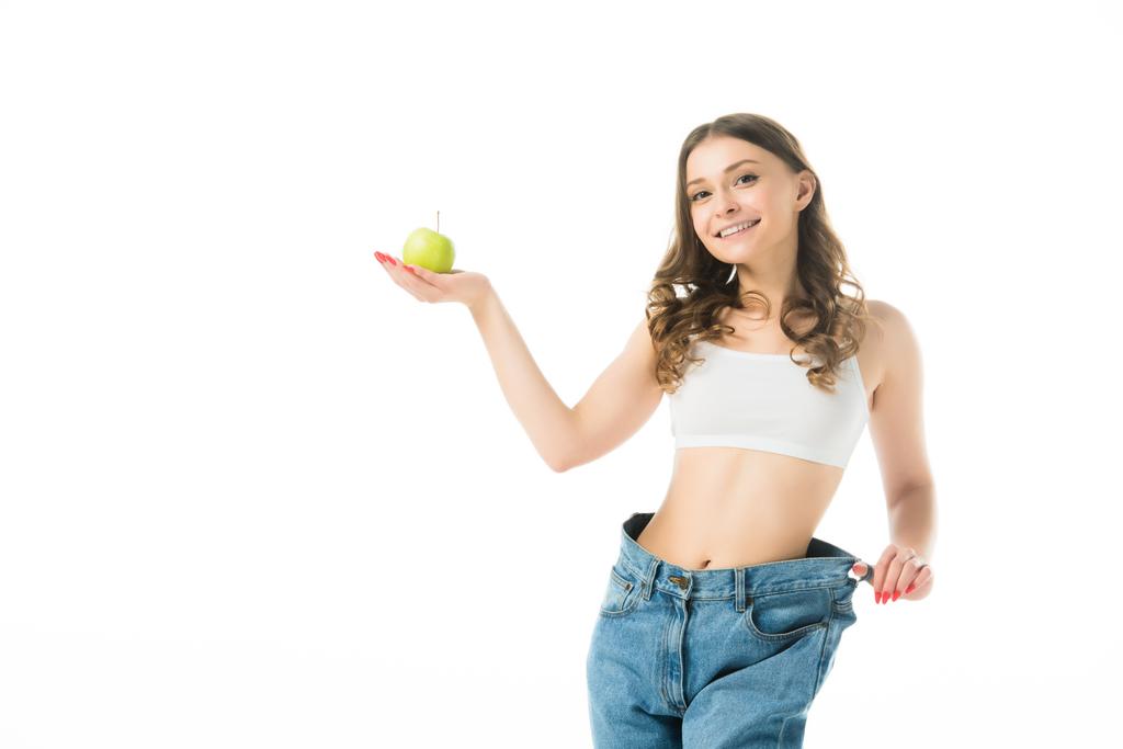 smiling slim young woman in big jeans holding green apple isolated on white - Photo, Image