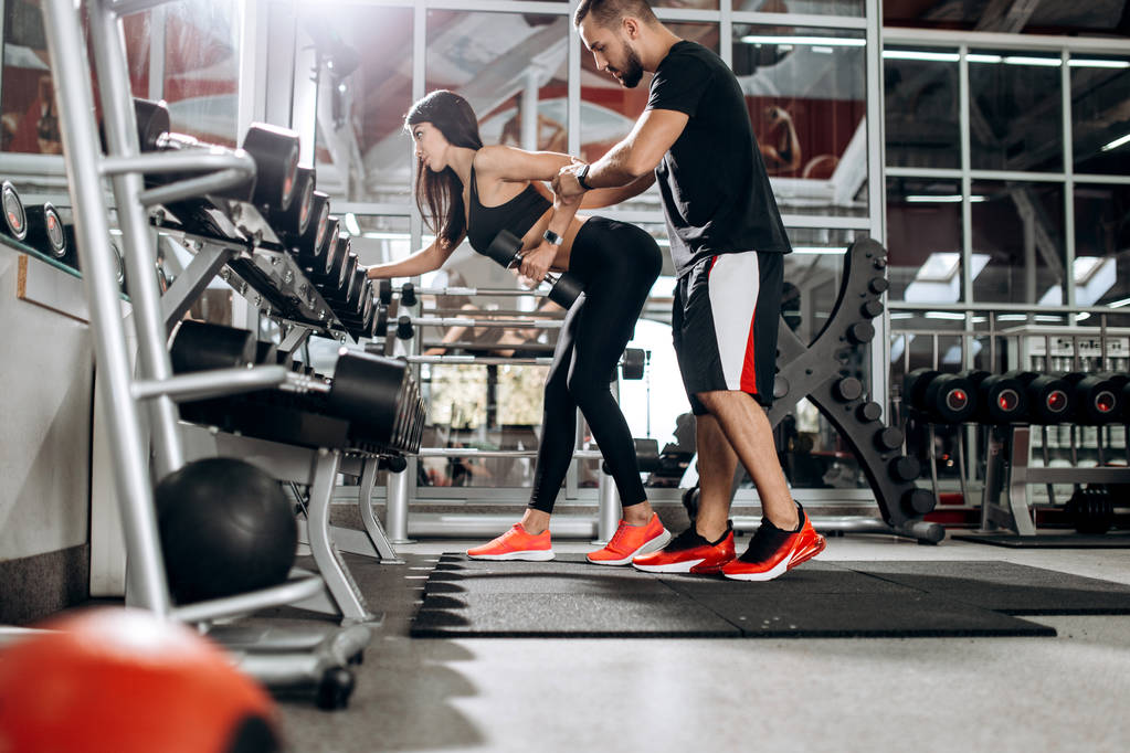 Slim κοπέλα ντυμένη στα μαύρα αθλητικά ενδύματα κάνει άσκηση τρικέφαλος μύς με αλτήρα με καναπέ βοήθεια στο γυμναστήριο - Φωτογραφία, εικόνα