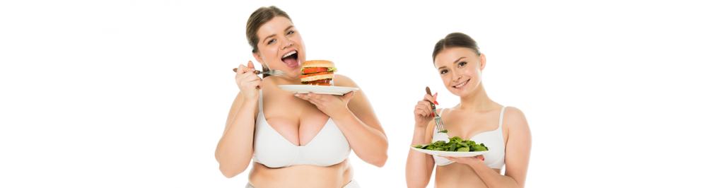 υπέρβαροι χαμογελαστό γυναίκα εσώρουχα τρώει burger από πλάκα ενώ slim ευτυχισμένη γυναίκα τρώει φύλλα πράσινο σπανάκι που απομονώνονται σε λευκό - Φωτογραφία, εικόνα