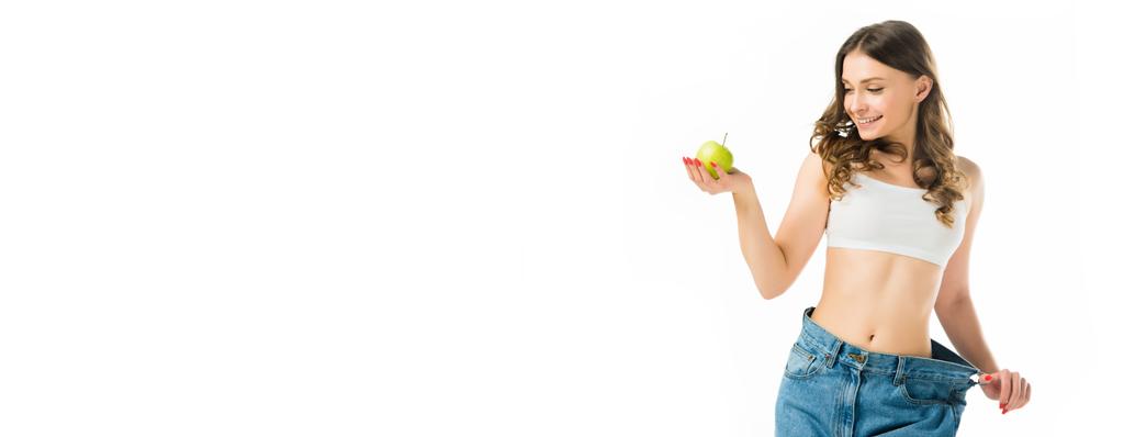 lachende slanke jonge vrouw in grote spijkerbroek bedrijf rijpe groene appel geïsoleerd op wit - Foto, afbeelding