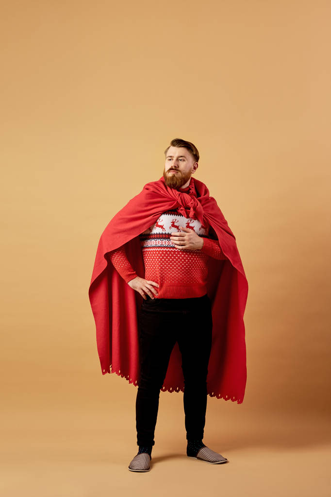 Red-haired man with beard dressed in a red and white sweater with deer and red cape and sleepers stands on a beige background - Photo, Image