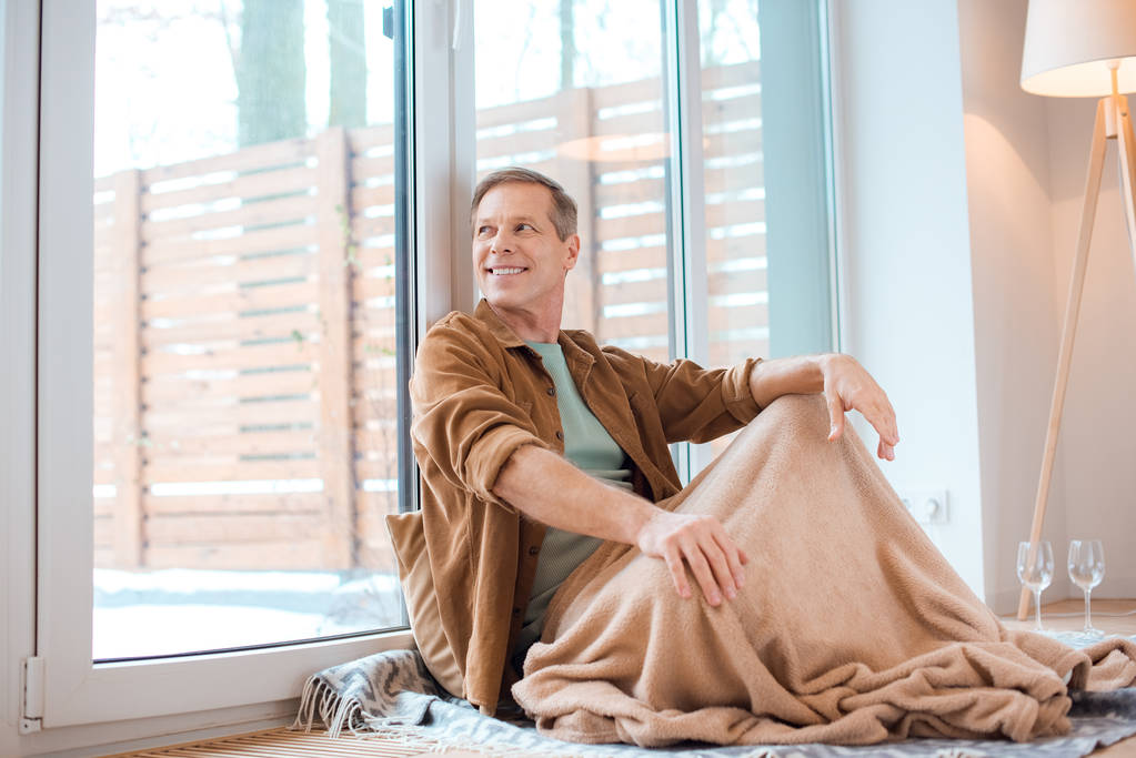 homem feliz sentado no chão sob cobertor de lã macia e olhando através de uma grande janela
 - Foto, Imagem
