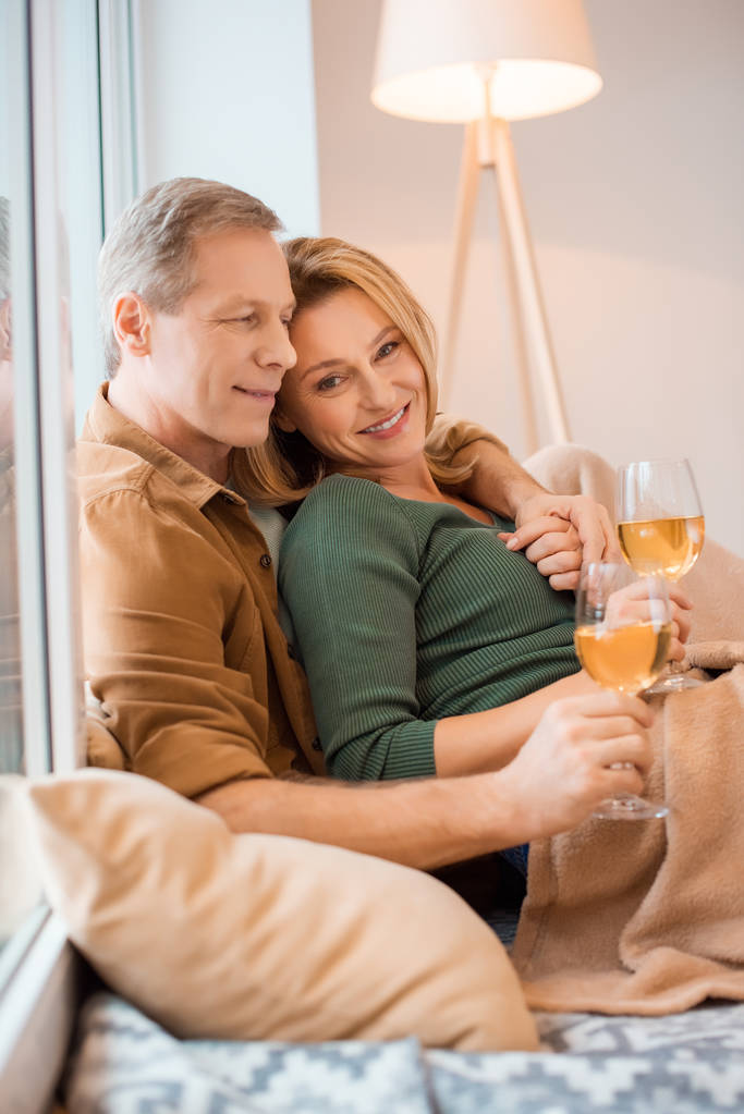 χαμογελώντας ζευγάρι αγκαλιάζει και κρατώντας ποτήρια λευκό κρασί, ενώ αναπαύεται στο πάτωμα στο νέο σπίτι - Φωτογραφία, εικόνα