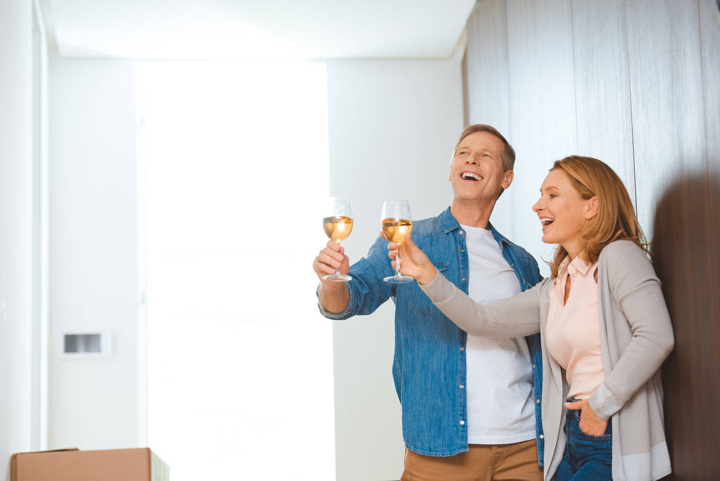 ενθουσιασμένος ζευγάρι κρατώντας ποτήρια λευκό κρασί ενώ στέκεται από τοίχο στο νέο σπίτι - Φωτογραφία, εικόνα