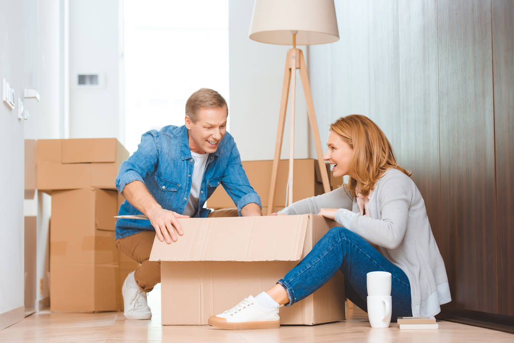 cheerful couple sitting on floor and unpacking cardboard box - Photo, Image