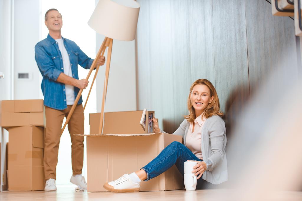 селективное внимание женщины, сидящей на полу и распаковывающей картонную коробку, в то время как муж держит напольную лампу
 - Фото, изображение