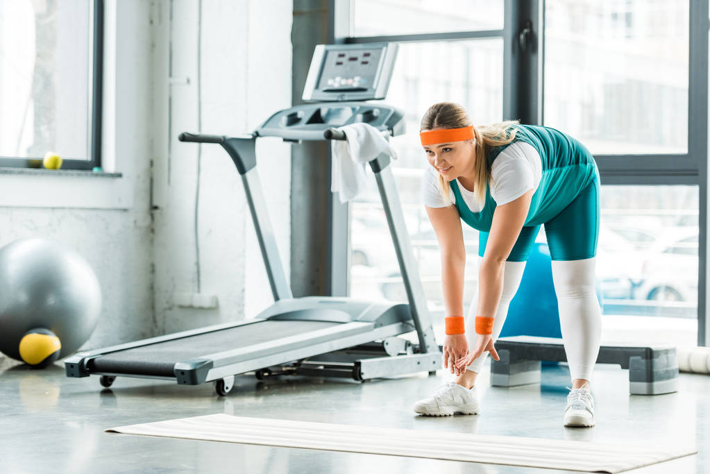 mujer con sobrepeso estiramiento cerca de la alfombra de fitness y la cinta de correr en el gimnasio
 - Foto, imagen