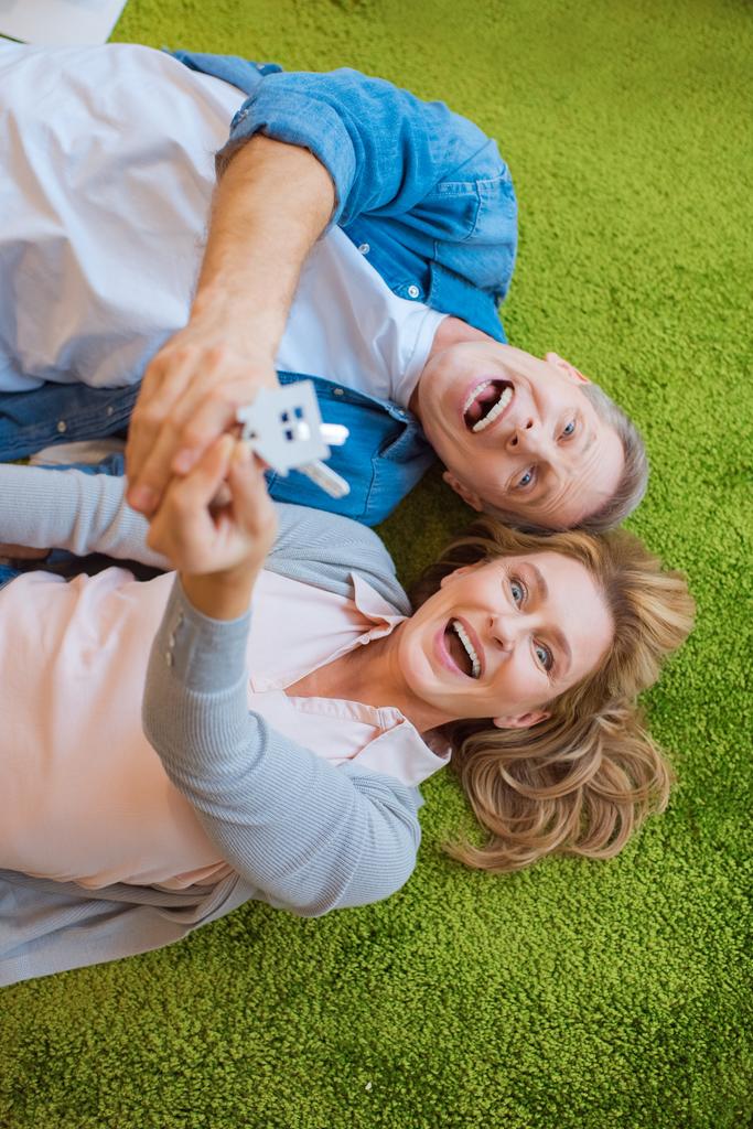 enfoque selectivo de pareja excitada sosteniendo llaves con baratija modelo de casa mientras está acostado en la alfombra verde
  - Foto, Imagen