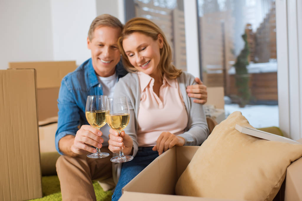 selective focus of happy couple clinking glasses of white wine while sitting by cardboard boxes at new home - Photo, Image