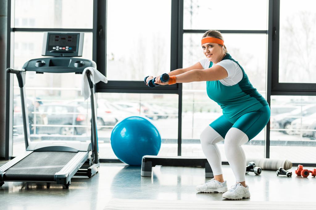 cheerful plus size woman exercising with dumbbells near treadmill  - Photo, Image
