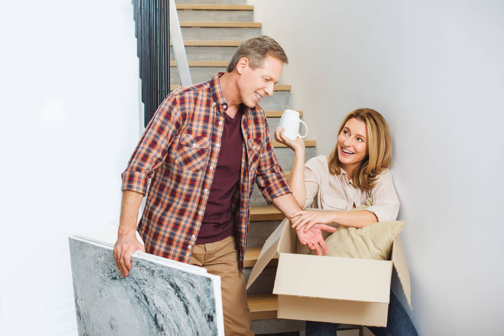 усміхнений чоловік тримає фотографію, стоячи біля дружини, сидячи на сходах і розпаковуючи картонну коробку
 - Фото, зображення