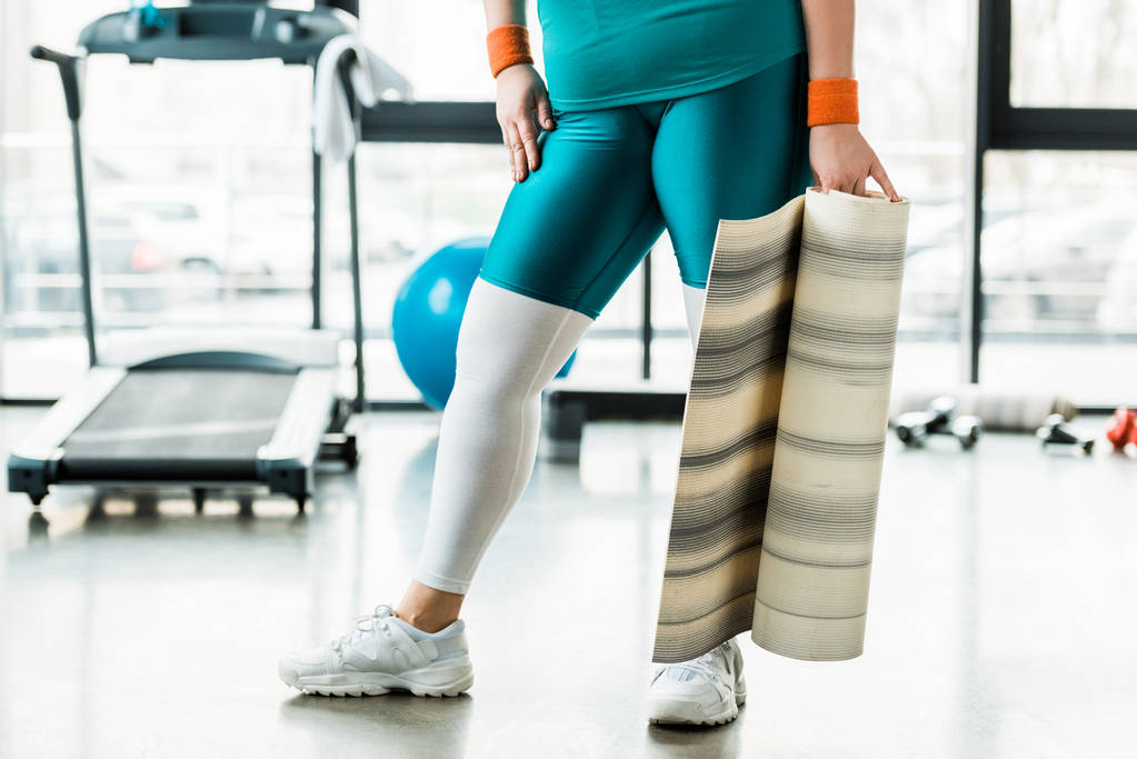 vista recortada de la mujer con sobrepeso que sostiene la estera de fitness en el gimnasio
 - Foto, imagen
