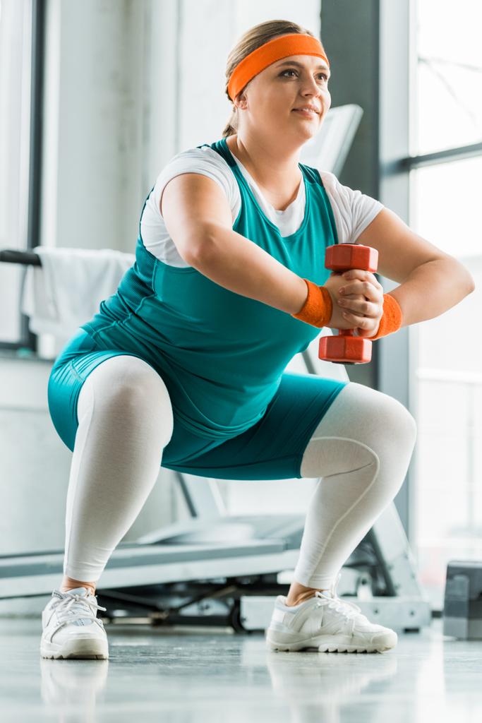 Lächelndes übergewichtiges Mädchen hockt mit Hantel im Fitnessstudio - Foto, Bild