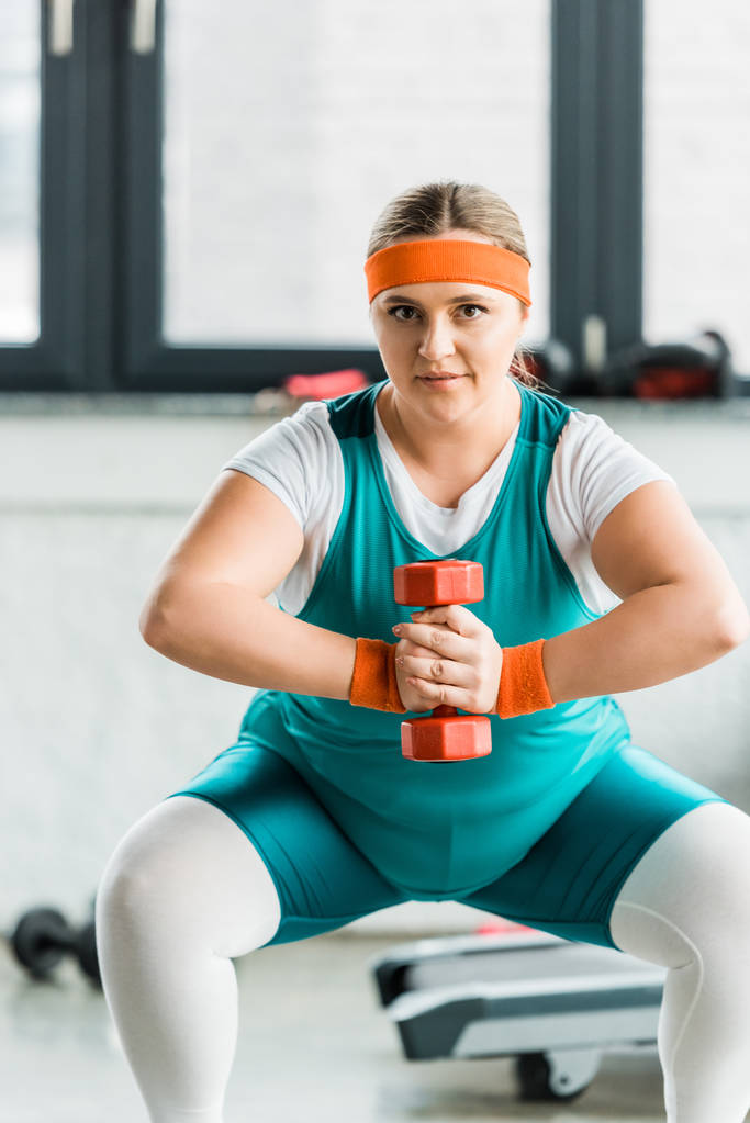 attraktives übergewichtiges Mädchen in Sportbekleidung hockt mit Hantel im Fitnessstudio - Foto, Bild