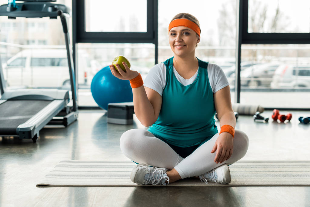 plus velikost ženy sedí na fitness mat poblíž sportovního vybavení se zkříženýma nohama a drží zelené jablko - Fotografie, Obrázek