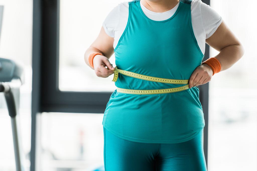 vista recortada de la niña con sobrepeso que mide la cintura en el gimnasio
 - Foto, Imagen