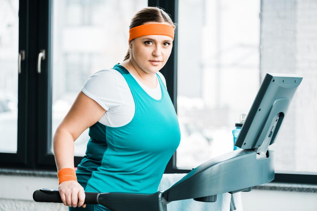 привлекательный избыточный вес девушка упражнения на беговой дорожке, глядя на камеру в тренажерном зале
 - Фото, изображение