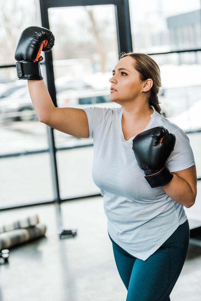  serious plus size woman wearing boxing gloves practicing kickboxing in gym - Photo, Image