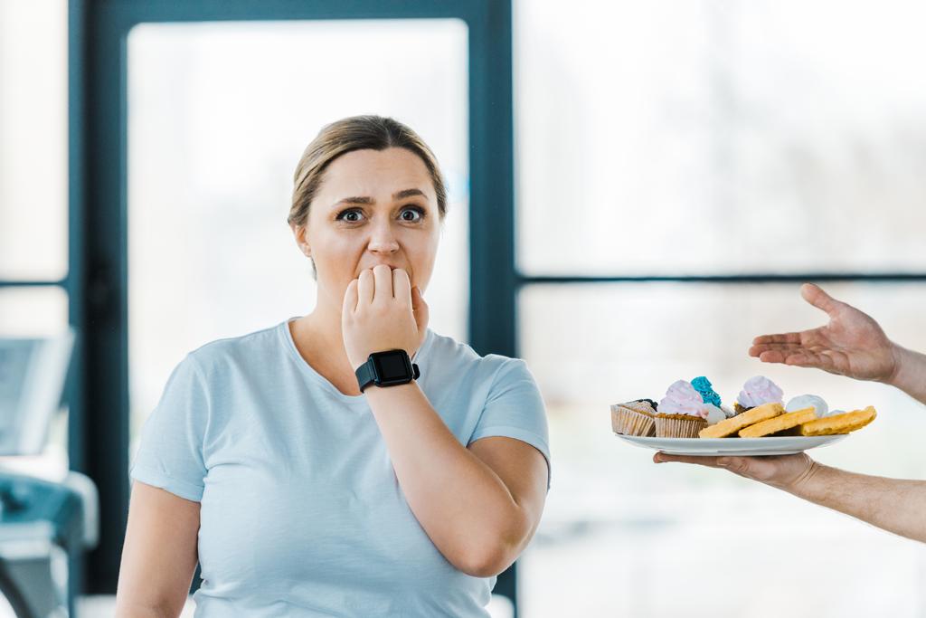 abgeschnittene Ansicht eines Mannes, der einen Teller mit süßem Gebäck in der Nähe einer übergewichtigen Frau hält, die den Mund in der Turnhalle bedeckt - Foto, Bild