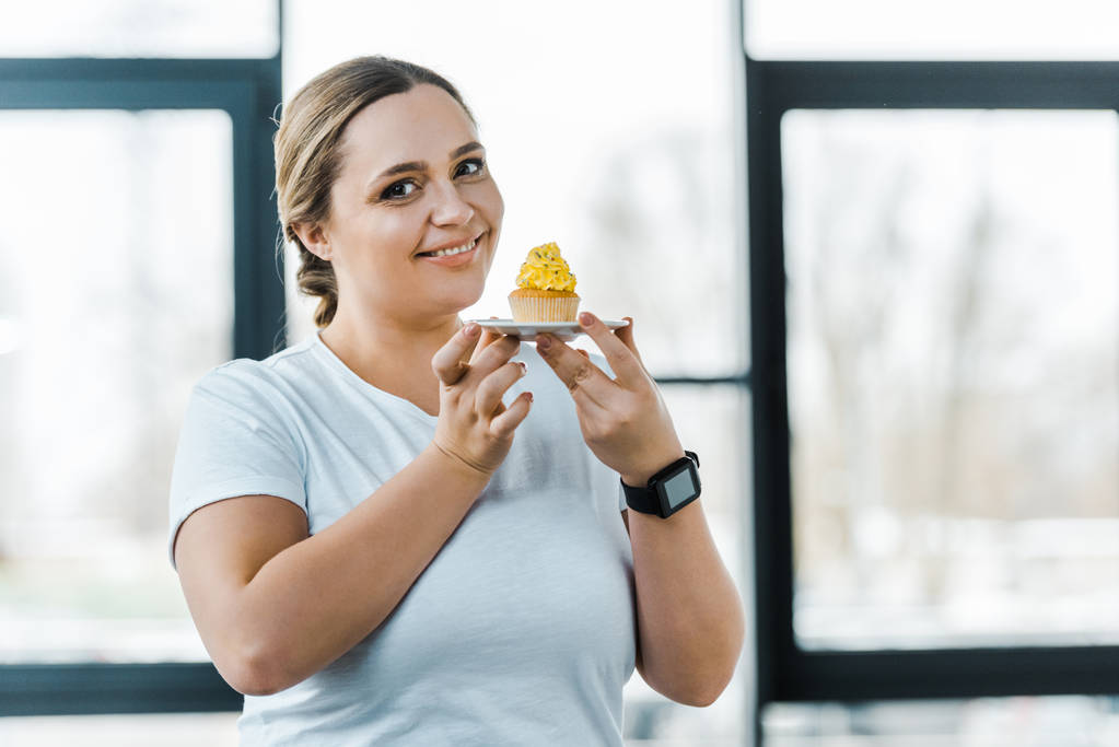 ευτυχισμένη υπέρβαρη γυναίκα κρατώντας νόστιμο cupcake στο γυμναστήριο - Φωτογραφία, εικόνα