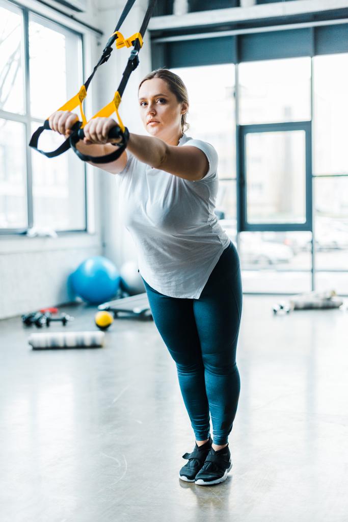 концентрированный избыточный вес девушки тренировки руки с подвеской ремни в тренажерном зале
 - Фото, изображение