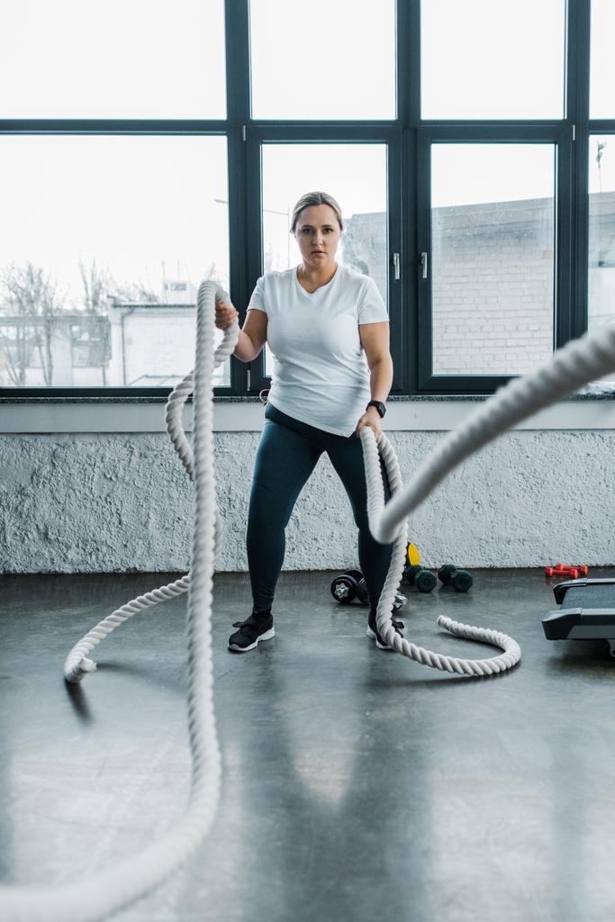 concentrated overweight woman training with battle ropes in gym - Photo, Image