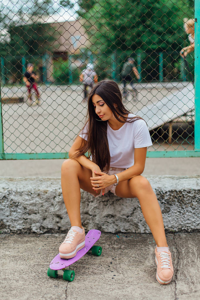 Sommer-Lifestyle-Bild des trendigen hübschen jungen Mädchens, das neben dem Skateboard-Wagen mit seinem Plastik-Skateboard sitzt. - Foto, Bild