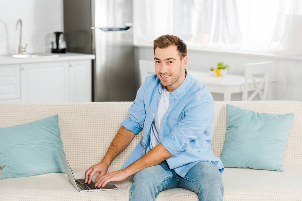 όμορφος άνδρας κάθεται στον καναπέ και βλέπουν φωτογραφική μηχανή χρησιμοποιώντας το φορητό υπολογιστή στο σπίτι - Φωτογραφία, εικόνα