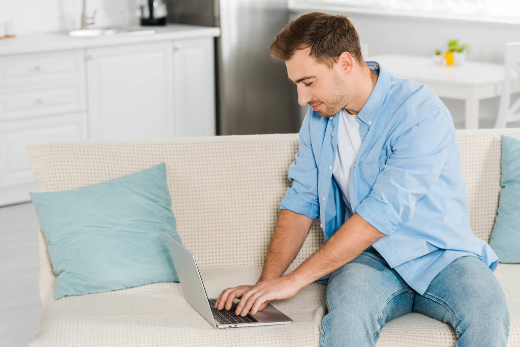 красивый мужчина, сидящий на диване и использующий ноутбук дома
 - Фото, изображение