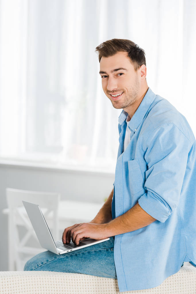 красивый улыбчивый мужчина сидит, смотрит на камеру и использует ноутбук дома с копировальным пространством
 - Фото, изображение